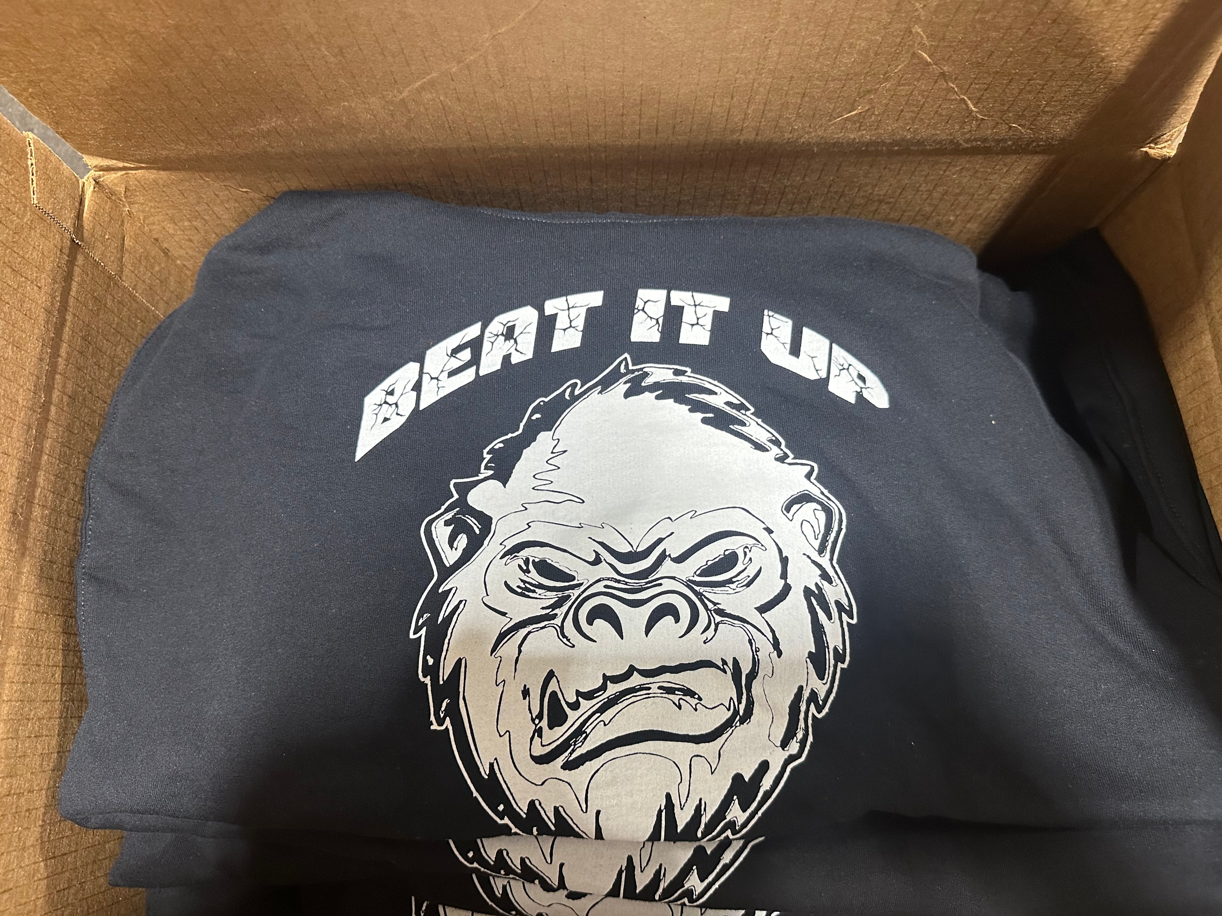 Black t-shirt with a gorilla graphic and 'Beat It Up' text in a cardboard box.