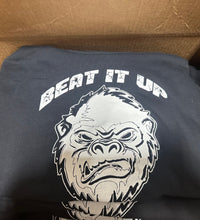 Black t-shirt with a gorilla graphic and 'Beat It Up' text in a cardboard box.